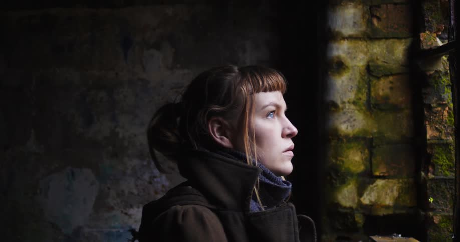 A white woman walks over to a window in a decrepit building and looks outside