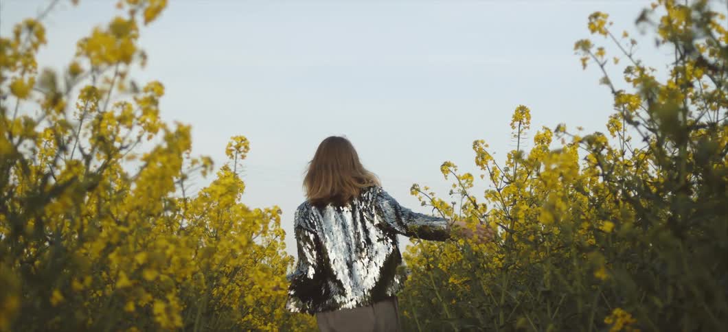 A woman in a sequined shirt walks through a field of yellow flowers