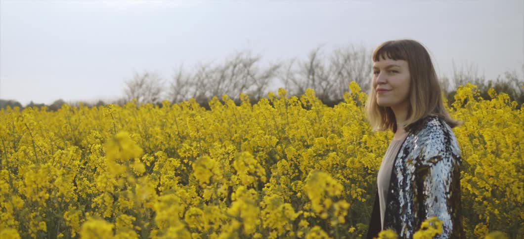Woman walking on a field of yellow flowers looks at the camera and smiles