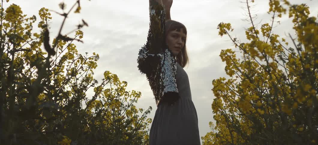 A white woman slightly smiles as she raises her arms and moves up and down in a field of flowers