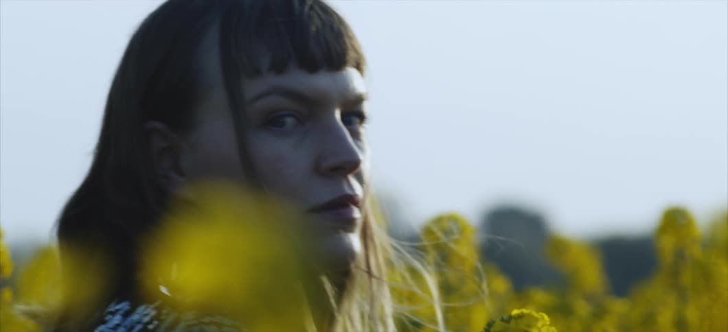 A woman walks through a yellow field looking at camera
