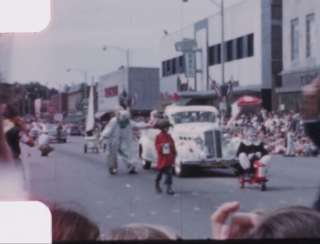 Vehicle leads a parade as clowns walk nearby and a crowd watches