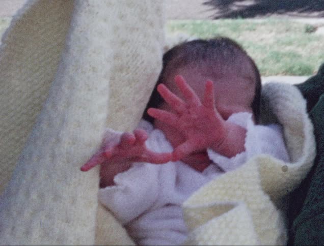 Person cradles young baby in blanket standing outdoors near lawn and street