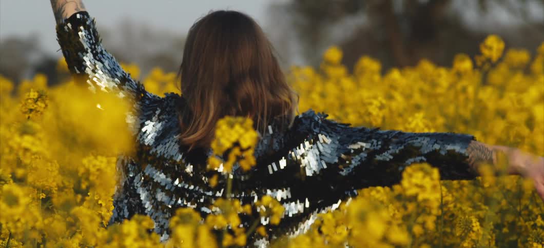 A woman wearing a glittering jacket walks through a field full of flowers