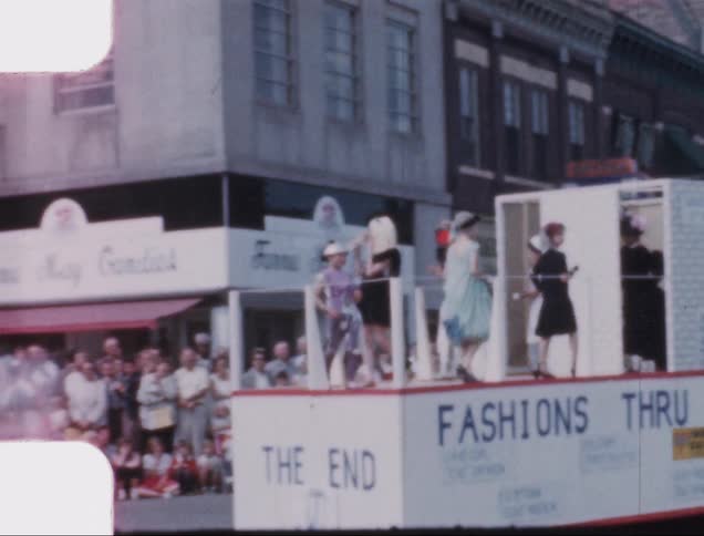 Women putting on a historical fashion show atop a parade float as the crowd watches it travel along the street