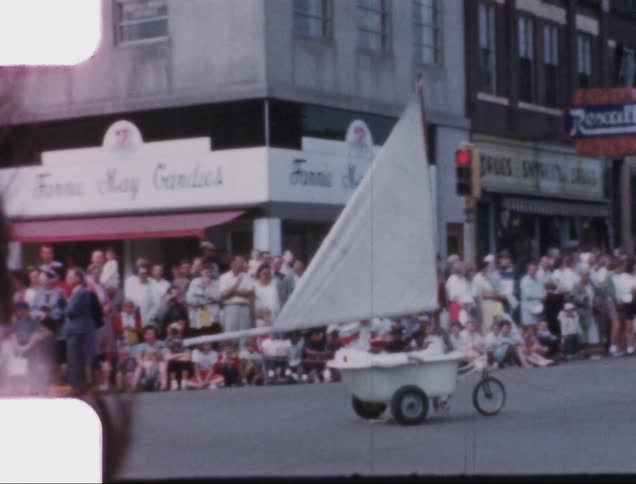 A clown riding down a street in a bathroom with a sail and wheels attached as part of a parade