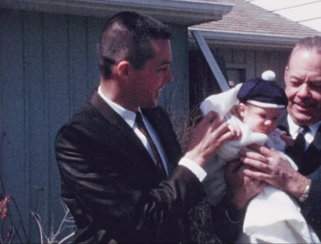 A grandfather holds his grandchild as the father looks on