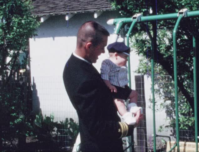 A man in a military uniform holds his son while his daughter plays on a swing