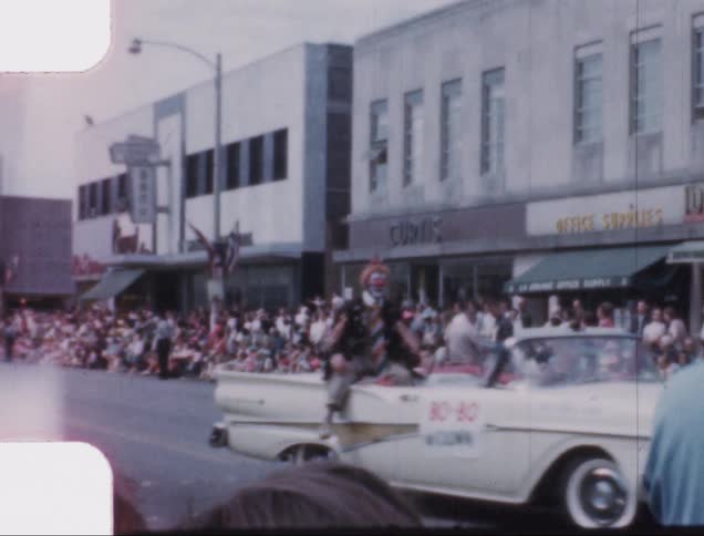Clown rides on back of white car along parade route waving to crowd
