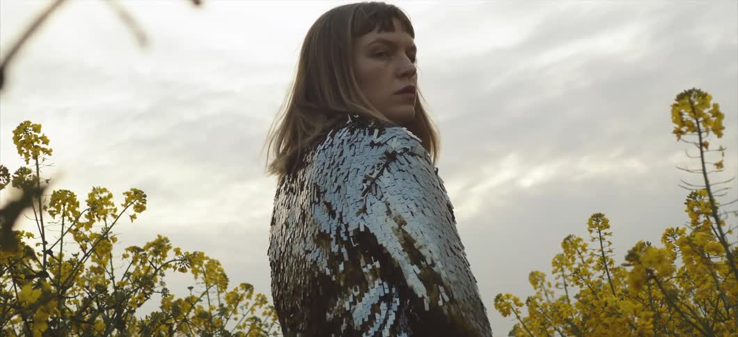 A woman standing wearing a silver blouse with plants and the sky in the background