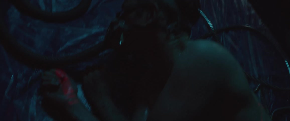 A man wearing a respirator attached to a long tube lying naked on a sheet of thin transparent plastic as red light is projected onto his skin in a dark room