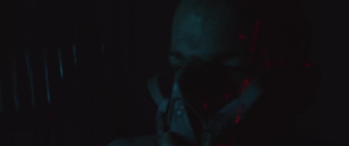 A white man wearing a mask sits in a dark room