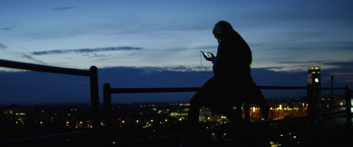 A woman sits and uses her cell phone on a balcony overlooking a city at night
