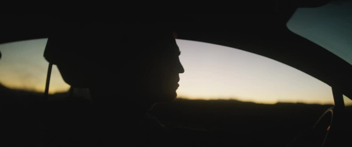 A silhouetted man sits in the driver seat of a car at dusk