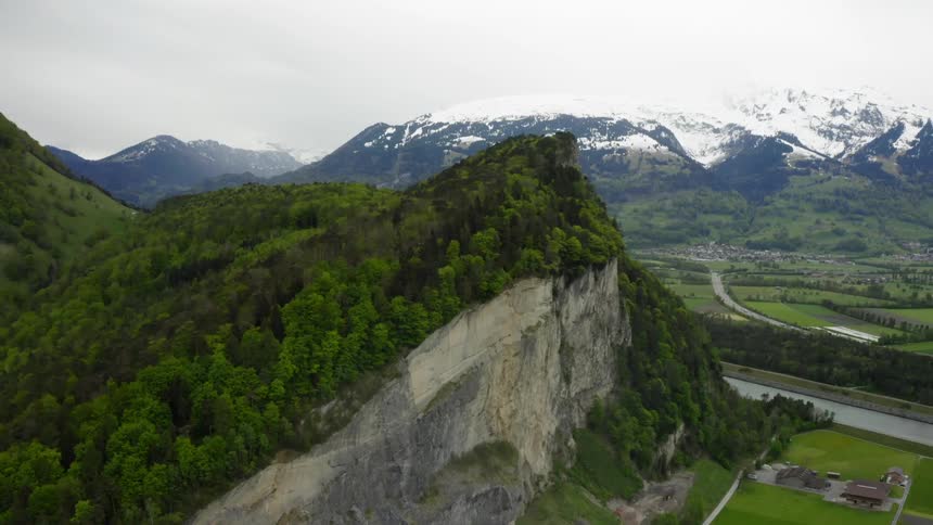 A mountain covered in trees with one side shaved off into a cliff rises above the terrain