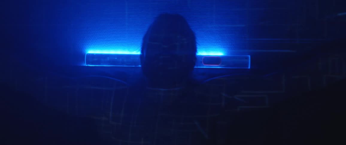 A shirtless man taking a seat in front of a blue light as geometric patterns are projected onto his body and the surfaces around him