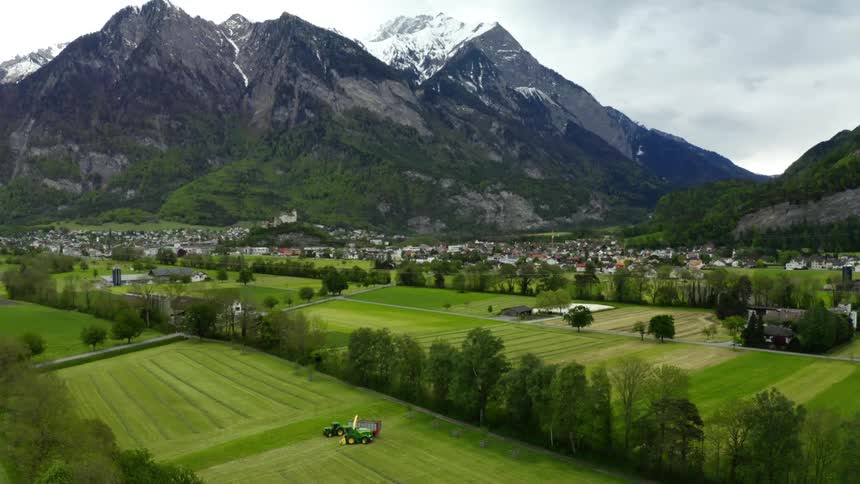 Harvest machine moves over field near town backed by mountains under an overcast sky