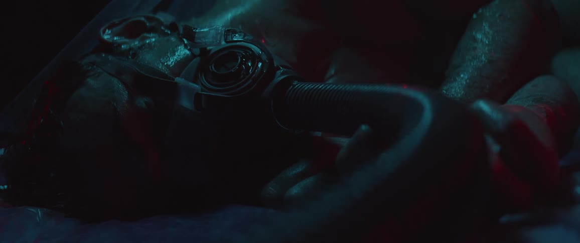 A white man lies in a dark room wearing a face mask