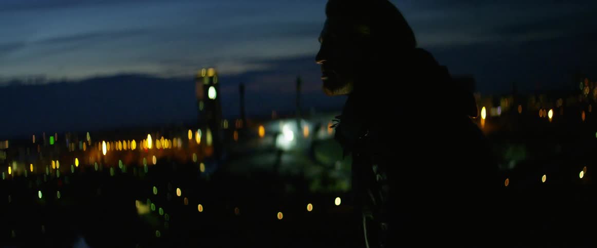 A man standing outdoor at high altitude in a city alternating between looking down at his smartphone and looking out over the city