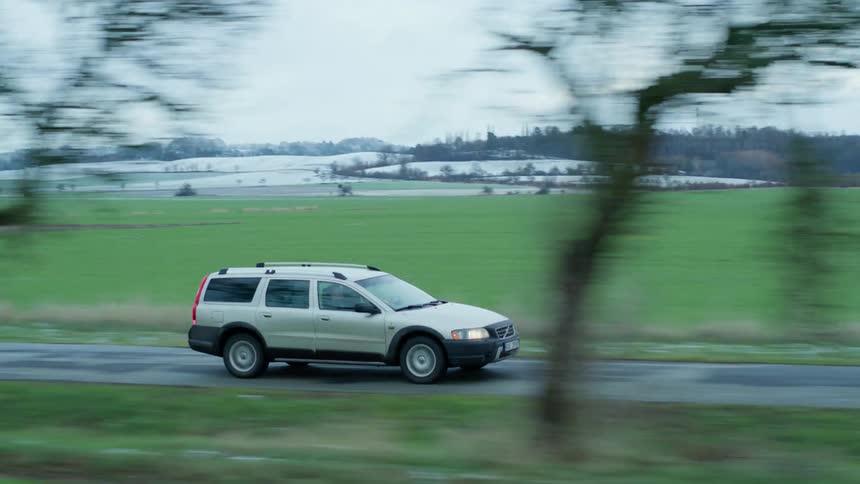 A vehicle driving on a road with a grassy field and buildings in the background