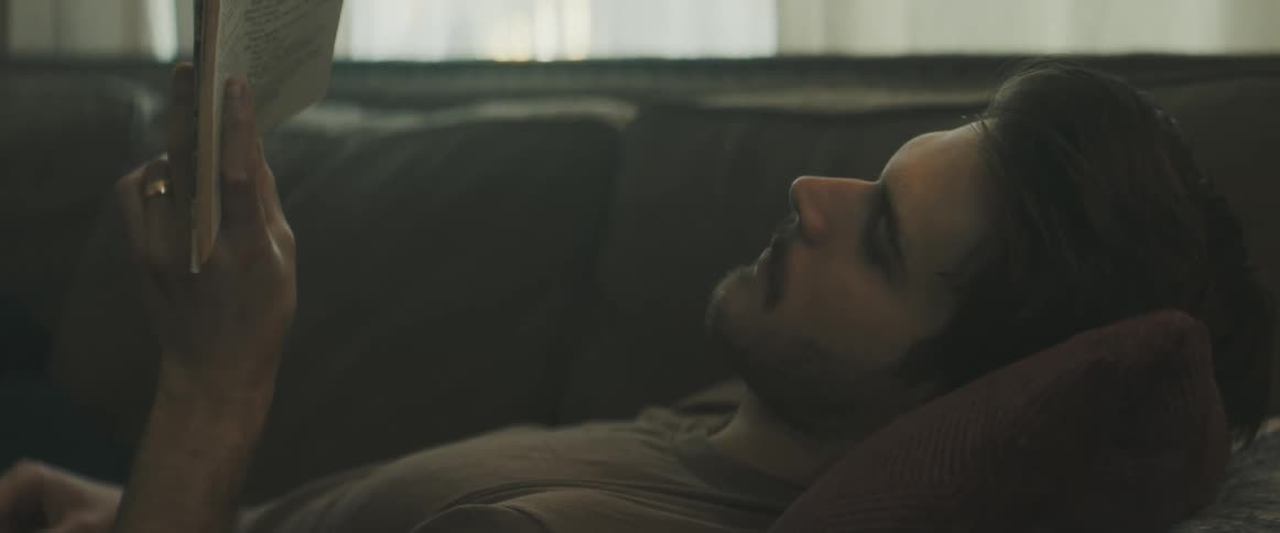 A man lies on the couch and reads and then a woman comes and sits by him