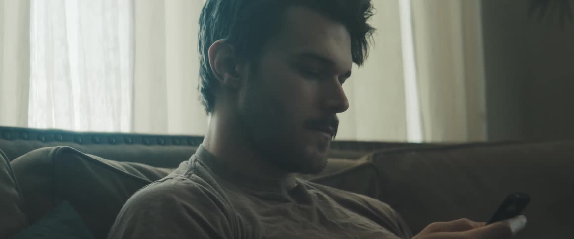 A man sits on his brown couch and scrolls through his phone