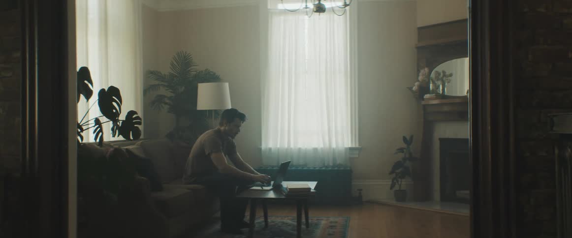 A white man sit on a couch in a living room typing on a laptop sitting on a coffee table