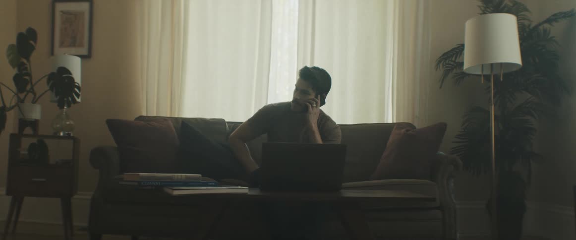 Male sitting on sofa at home typing on laptop puts laptop on coffee table to answer the phone and then stands up to walk out of the room