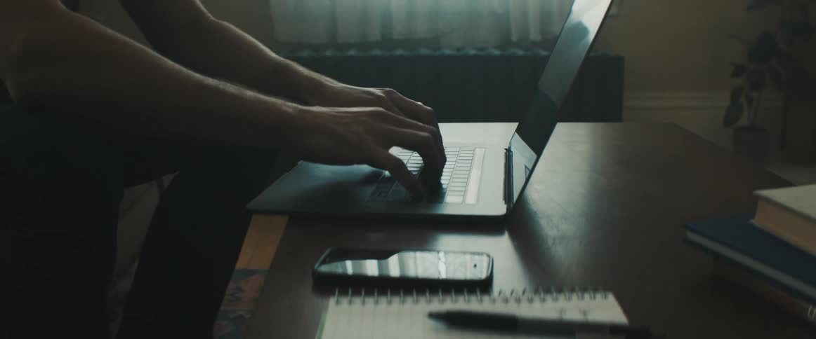Man types on a laptop while a cell phone sits nearby