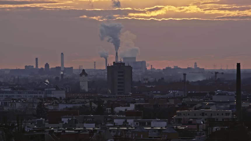 Smoke billows out of factories in a dense neighborhood surrounded by apartment buildings at sunset