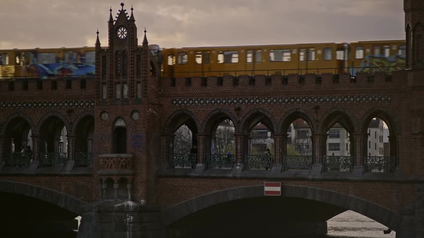 A train passes above a pedestrian bridge over a river in a city