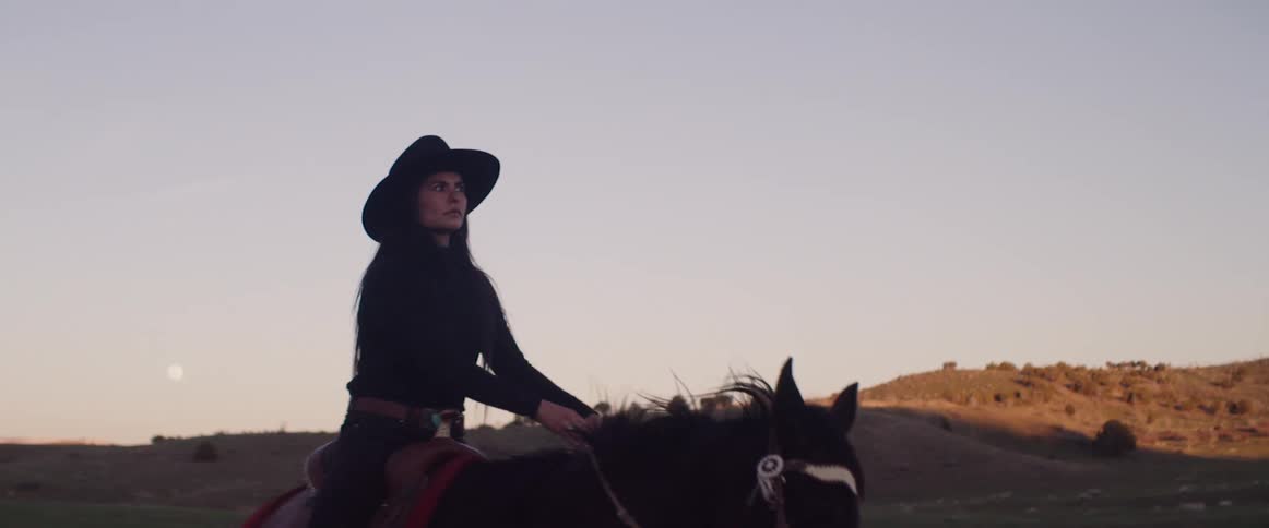 A woman rides a horse through a ranch near a hillside