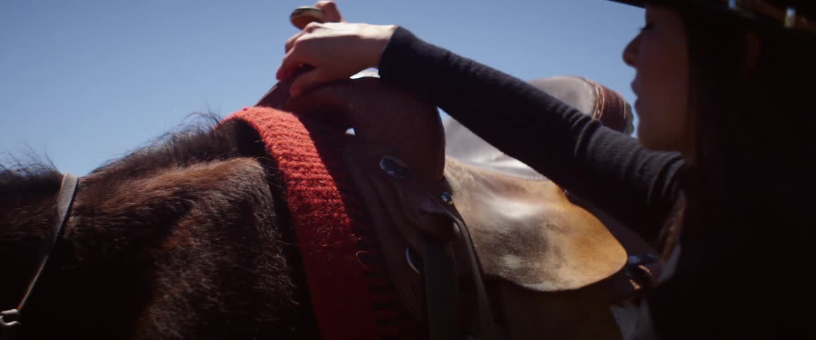 A woman in a black cowboy hat adjusting the saddle of a horse on a sunny day