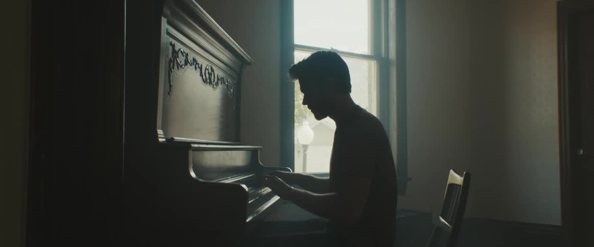 A man sits alone in a dim room by a window playing the piano
