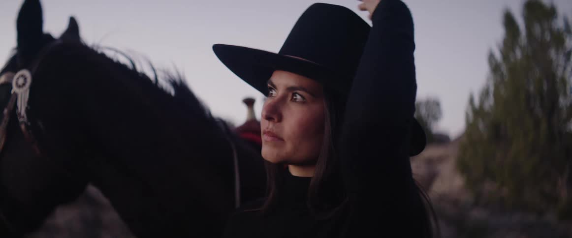 Woman standing by a dark horse wears a wide hat