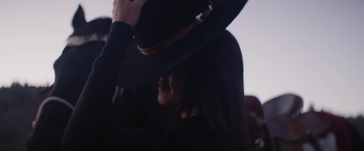 A white woman puts on a hat while standing next to her horse out in nature