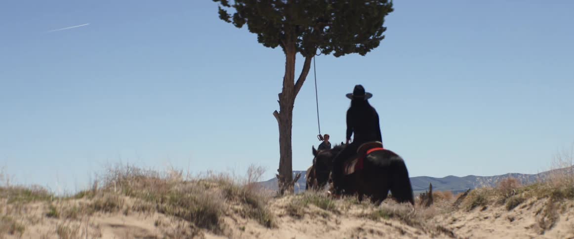 A person riding a horse with a person with a noose around their neck sitting on a horse underneath a tree