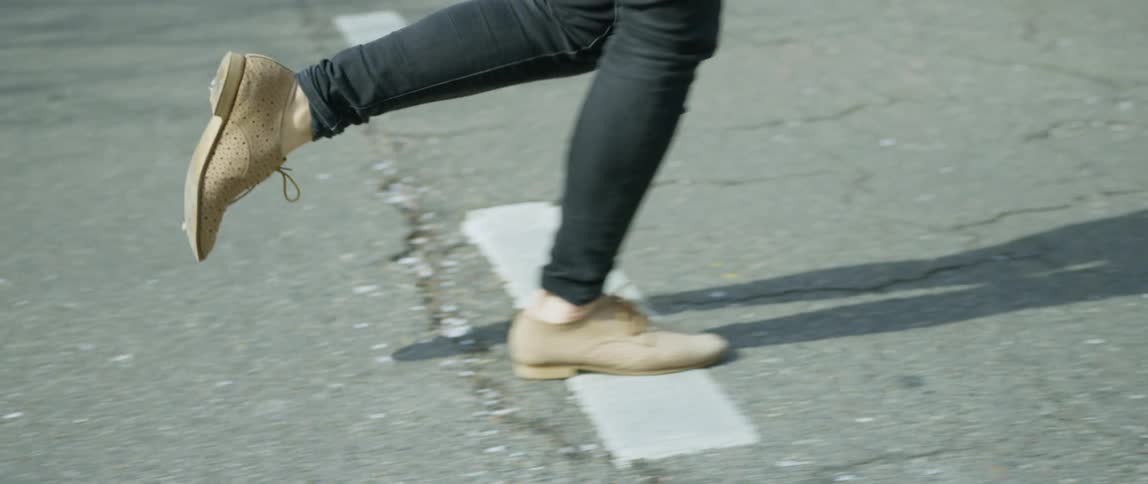 A woman wearing oxford shoes and black jeans runs across a street