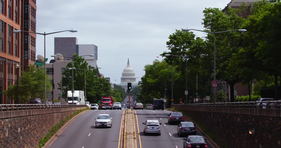 Vehicles travel along a city road beside trees and large buildings