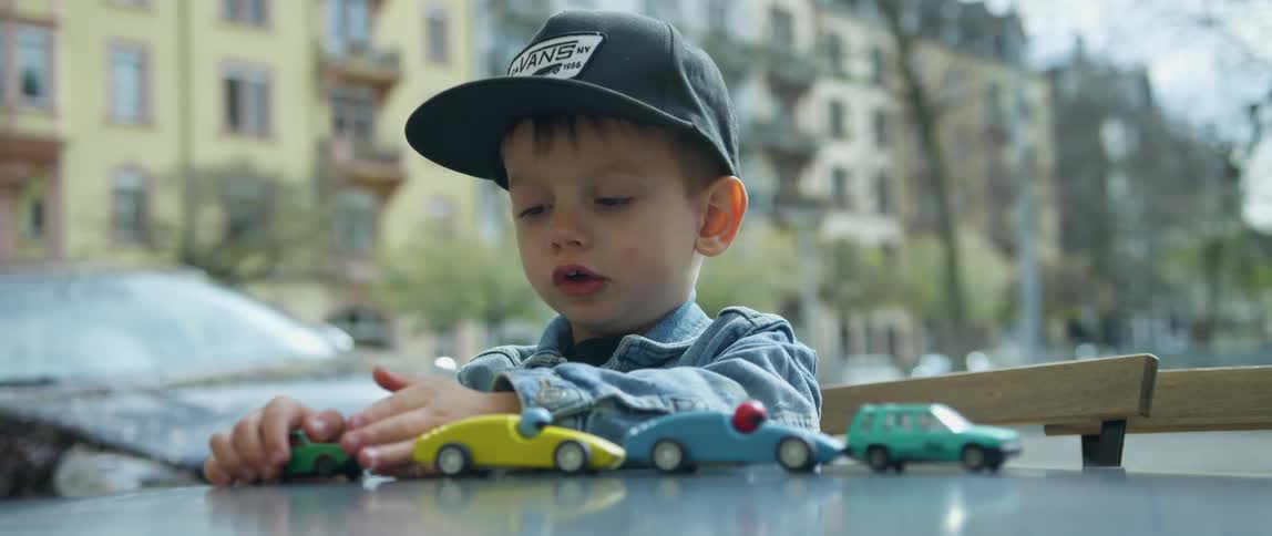 A little white boy plays with his toy cars outdoors in a city neighborhood