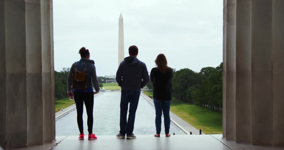 Three people stand and look at a monument and a woman takes a picture of it with her phone