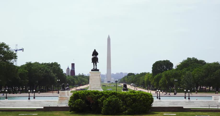 Horseman statue stands on pedestal near pond with washington monument and city in background as pedestrians walk around