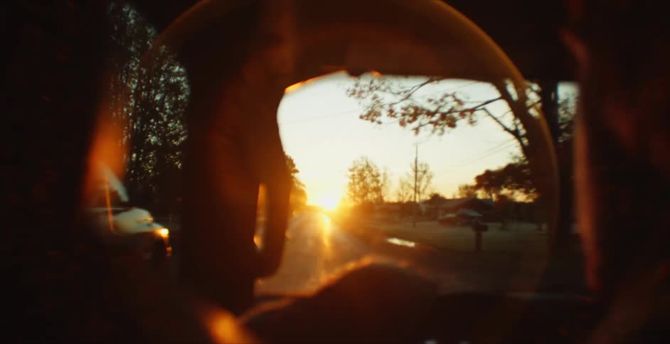 Man and woman drive along road past country homes toward sun on the horizon