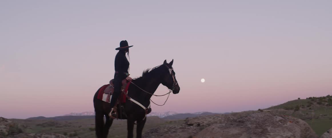 A cowgirl rides a horse in a rural area at dusk