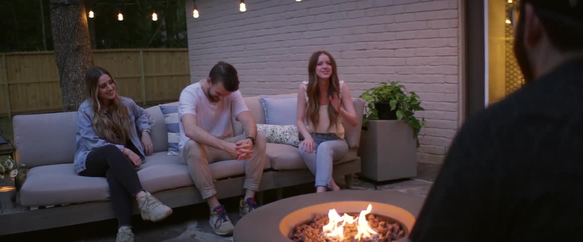 A group of laughing friends sits around a fire pit in a back yard