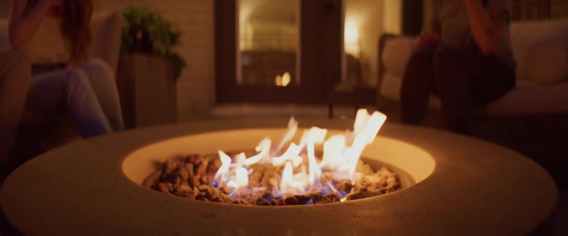 People sit on a furniture on a patio as a fire burns in a fire pit