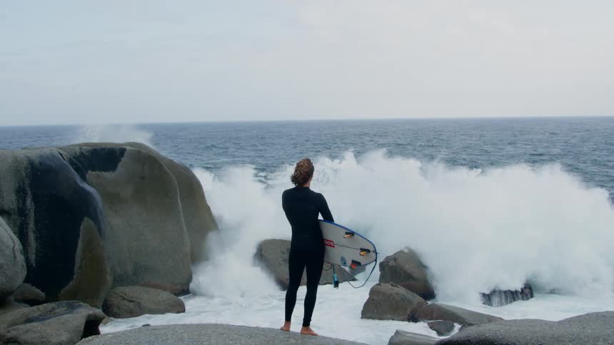 A surfer hold a board stands on the shore and watches a big wave come in