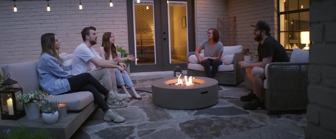 Five friends sit on a patio around a fire pit talking and laughing