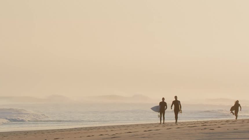Surfers carry surfboards across beach under cloudy slightly pink sky as waves roll in