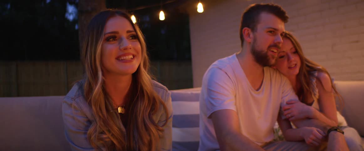A couple and a woman sit outdoors on a patio laughing as they hold sticks pointed to a fire
